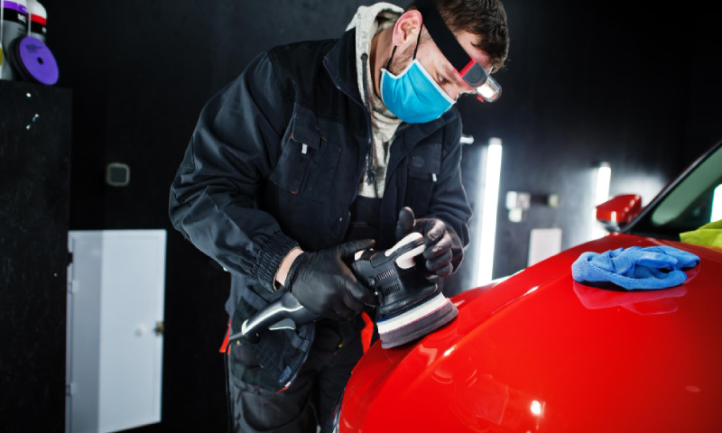 Car detailing concept Man in face mask with orbital polisher in repair shop polishing orange suv car<br />