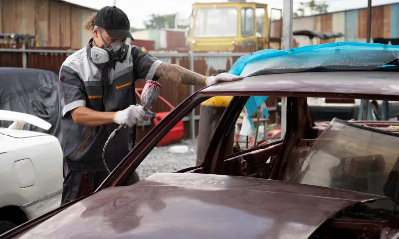 A mechanic in protective gear uses a spray gun to paint the roof of a car in a busy auto repair shop with various vehicles around