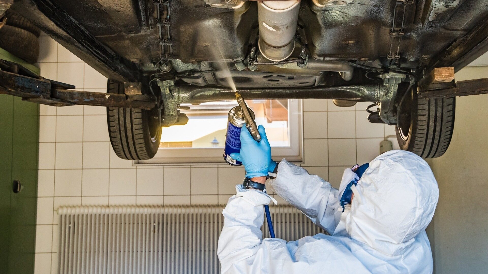 A worker in protective gear spray paints the side of a car in a bright, well-lit paint booth, with protective coverings in place