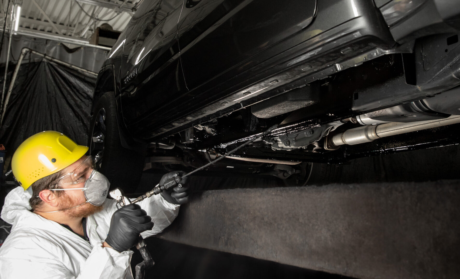 A worker in a black shirt and gloves inspects a blue car's rear bumper closely in a well-lit garage environment